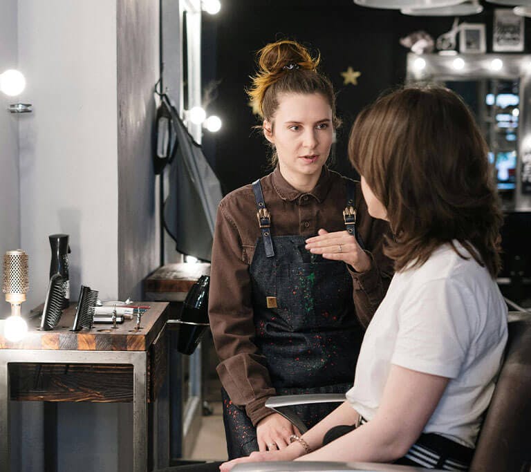 A person wearing an apron talking with another person seated in a salon chair, surrounded by hair styling tools and mirrors in a well-lit salon.