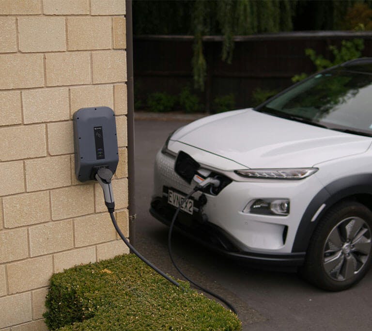 Electric vehicle parked beside a building and plugged into a wall-mounted charging unit with a cable running to the front of the car.