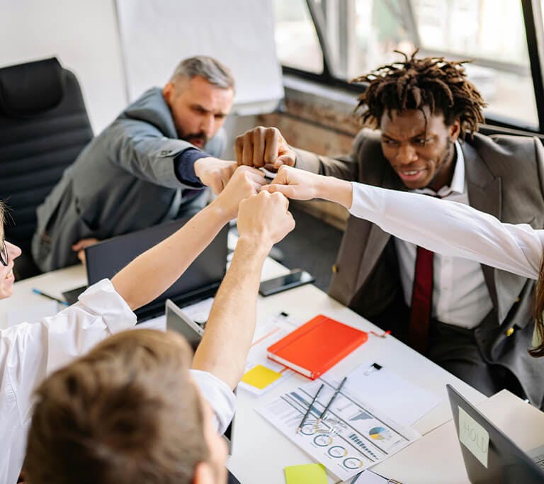 A group of colleagues sitting around a conference table enthusiastically fist-bumping in a show of teamwork and celebration during a meeting.