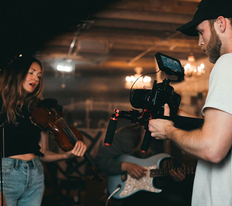 A videographer films a musician playing a violin in a dimly lit studio, with another musician playing guitar in the background.