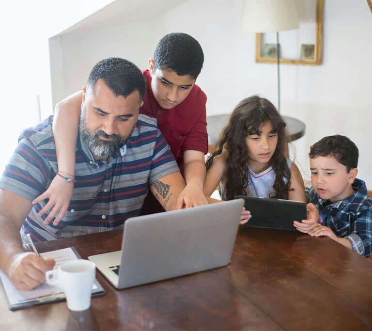 A parent and three kids are gathered around a table at home, working on a laptop and using a tablet.