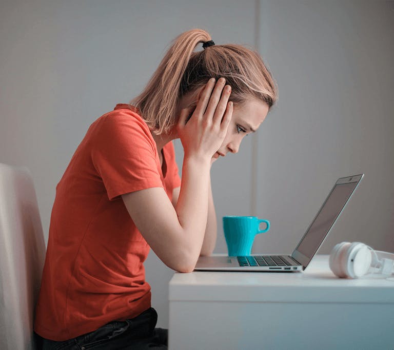 Person sitting at a desk, leaning toward a laptop with hands on their head, appearing frustrated or stressed. A turquoise mug and white headphones rest on the desk nearby.