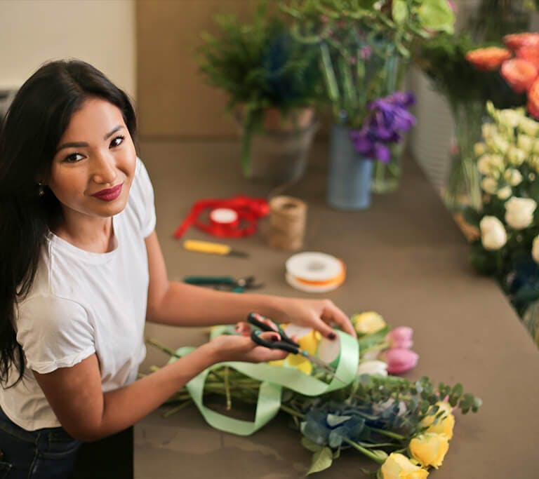 A person arranges a bouquet at a workstation, holding scissors and ribbon, with fresh flowers and floral tools spread across the table.