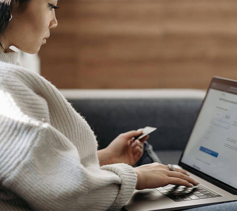 A person in an oversized, beige sweater holds a credit card in one hand and types on their laptop with the other.