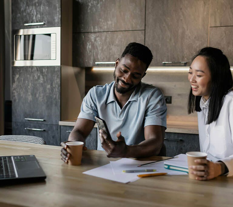 A couple stands at a kitchen island in front of a laptop and several papers and writing utensils as they drink coffee and smile at a phone.