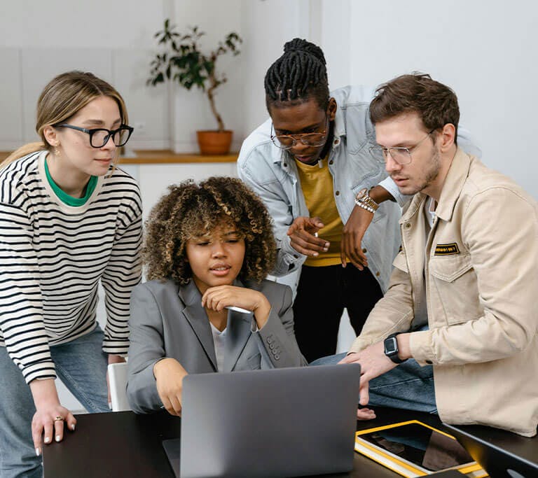 Four people gather around a laptop in a modern office setting, collaborating on a project. One person is seated and using the laptop while the others lean in and discuss, indicating a team meeting or group brainstorming session.