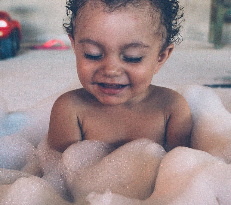 A baby with curly hair smiles in a bubble bath.