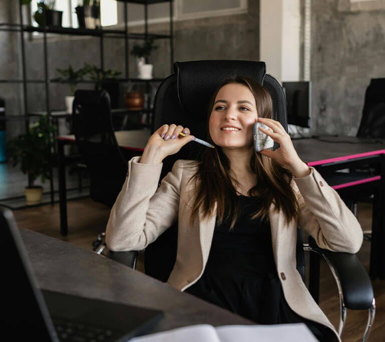 A person sitting at a desk in a modern office, smiling while talking on a mobile phone. They are leaning back in a chair and holding a pen, with a laptop and open notebook in front of them