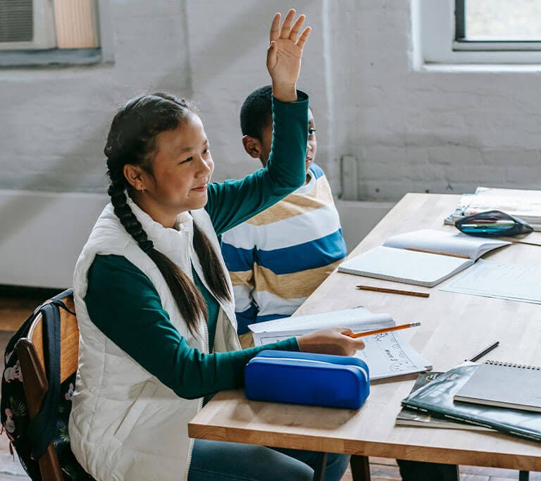 Two young students are sitting in a classroom, and one raises their hand to ask a question.
