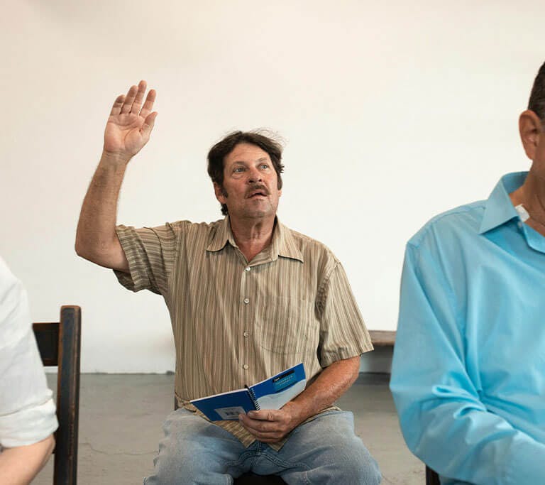 A person wearing denim jeans and a brown vertically striped shirt sits in a chair and raises their hand in a classroom with blank white walls.