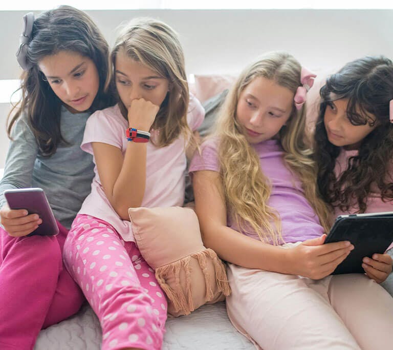 A group of preteens reclines on a bed while using their mobile devices, including a smartphone and tablet.