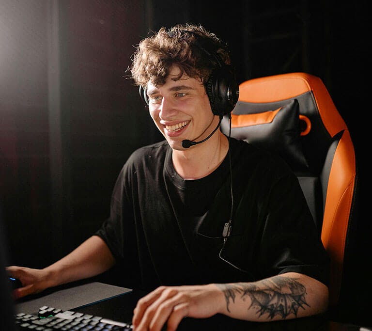 A person wearing a headset and sitting in an orange-and-black gaming chair smiles while playing on a desktop computer. They are using a keyboard and mouse in a dimly lit gaming setup.