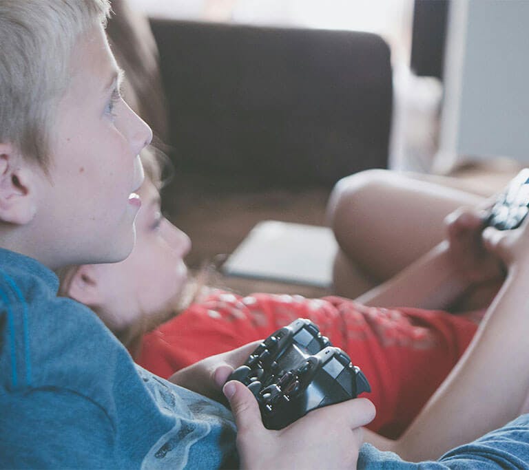 Two children sit comfortably and appear immersed as they play video games using PlayStation controllers.