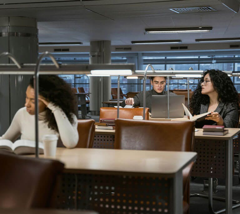 People studying quietly in a library or study area with books, laptops, and coffee cups on the desks. The space has modern, energy-efficient lighting, large windows and brown leather chairs arranged in rows.