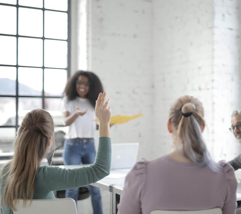 A group of people in a bright meeting room with large windows, engaged in a discussion. One person stands at the front speaking and smiling, while another raises a hand, indicating participation or asking a question.