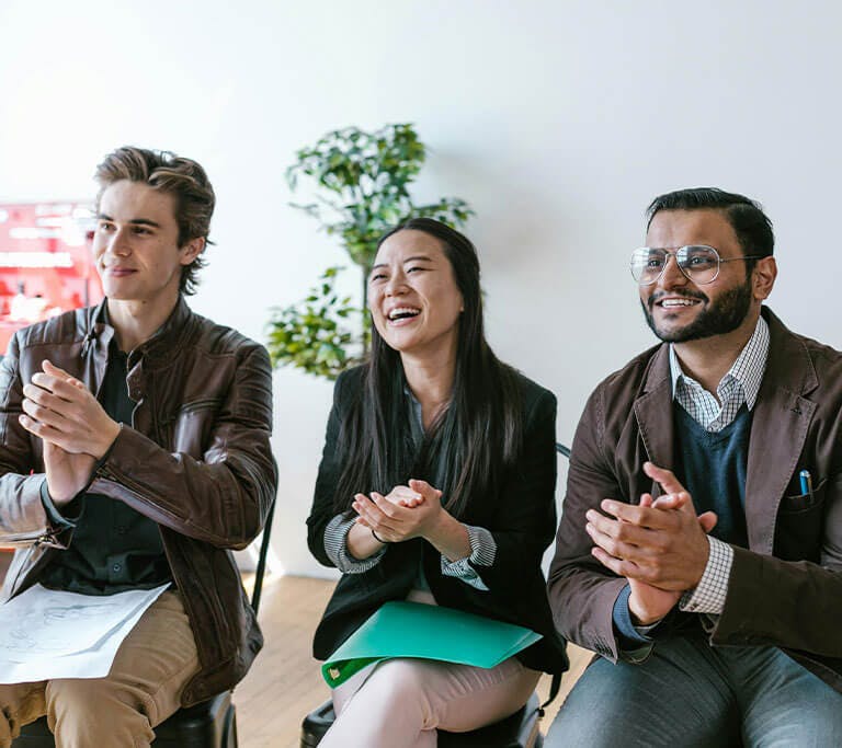 Three people sitting side by side in a bright office clap and smile during a presentation or meeting.
