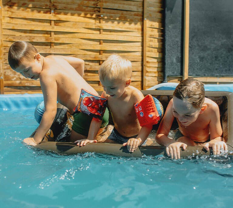 Three children wearing swim floaties play together on an inflatable raft in a backyard swimming pool on a sunny day.