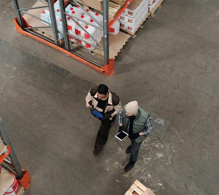 Two workers walking through a warehouse aisle, one holding a tablet and the other a clipboard, surrounded by shelves stocked with large containers and boxes.