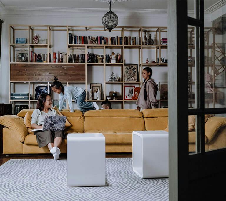 A smiling parent uses a laptop on a couch while three kids play and climb on the couch. They’re in a living room with a large shelving unit in the background, stocked with books and decorations.