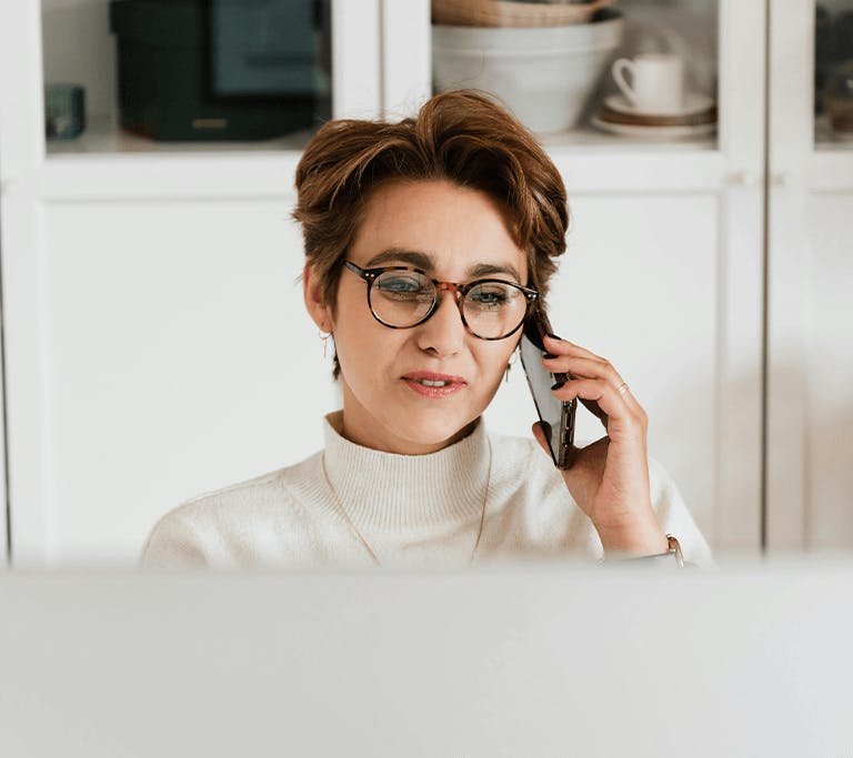 Person wearing glasses and a light turtleneck sweater speaks on a smartphone while sitting at a desk, partially obscured by a computer screen. Shelves with dishes and decor are visible in the background.