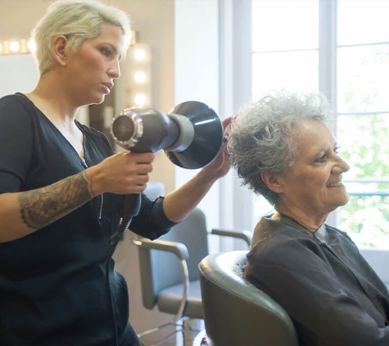 A hairstylist using a blow dryer to style a client’s short hair in a bright salon. Natural light filters through large windows and illuminated mirrors add a warm glow to the modern space.