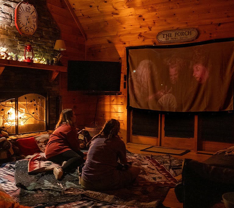 Two kids rest on quilts in a log cabin while streaming movies on a projector screen by the fire.