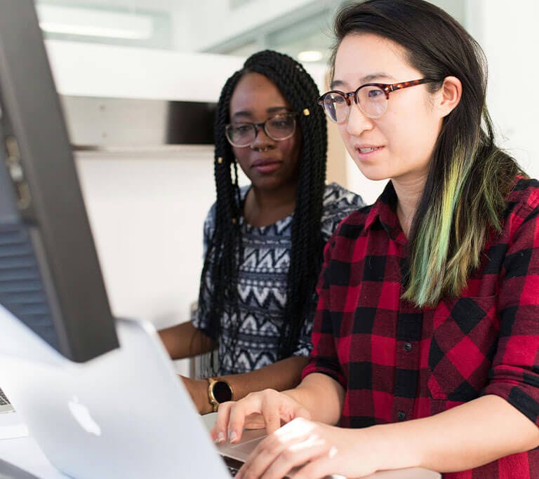 Two people work together at a computer in a bright, modern office. One is typing while the other observes the screen, suggesting collaboration or a learning session.