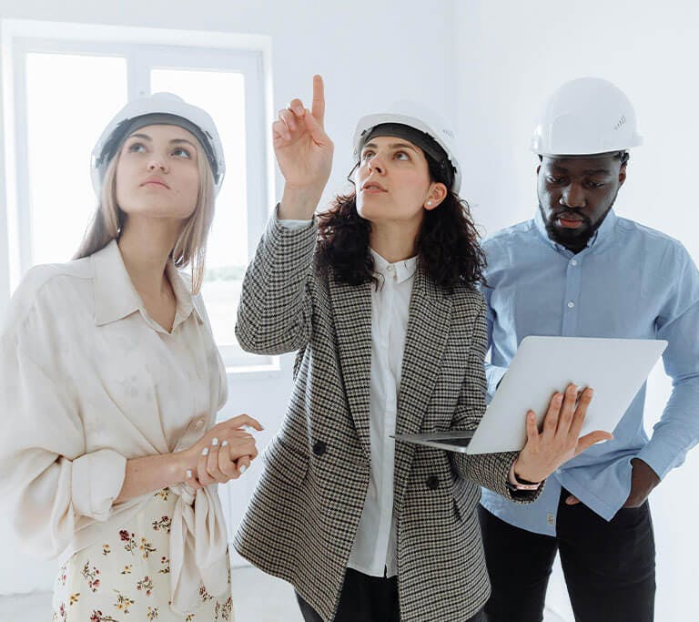 Three people wearing hard hats stand inside a bright, unfinished room. One person is pointing upward while holding a laptop, as the others look in the same direction, appearing to assess or discuss something on the ceiling.