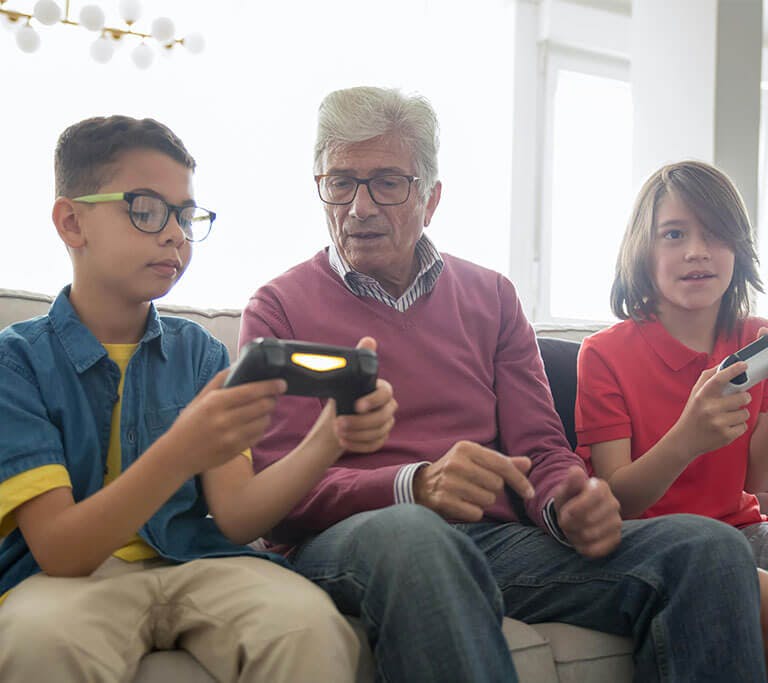 An older adult wearing a collared shirt under a mauve sweater and jeans sits on a couch next to two children who are holding video game controllers as one child demonstrates how to use the controller.