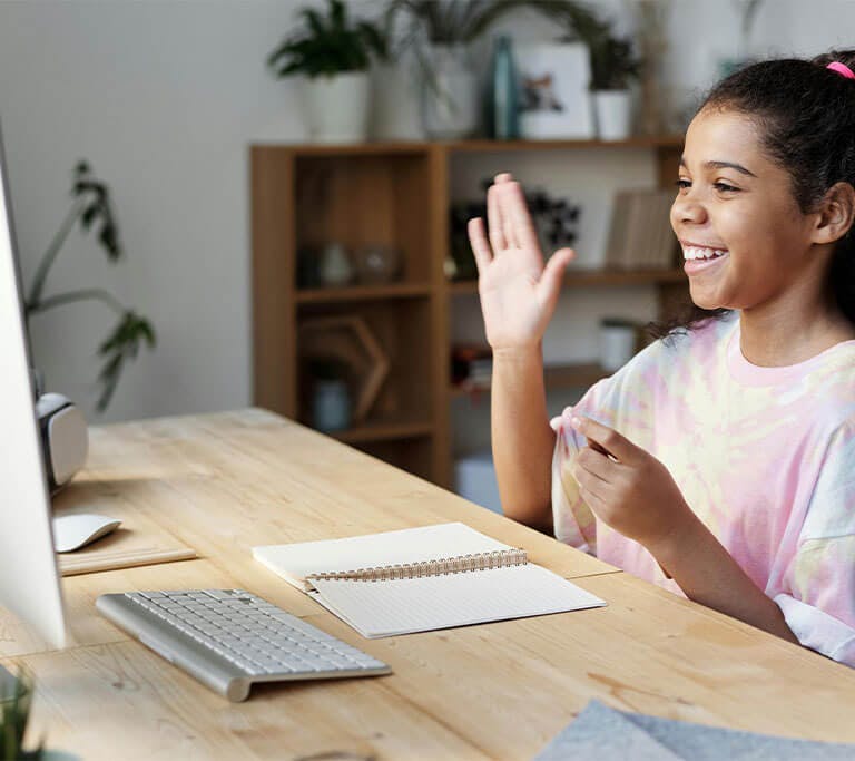 A student wearing a pastel tie-dyed shirt smiles and raises one hand, as if to ask a question, while sitting at a desk in front of a computer screen, mouse, keyboard and open spiral-bound notebook.