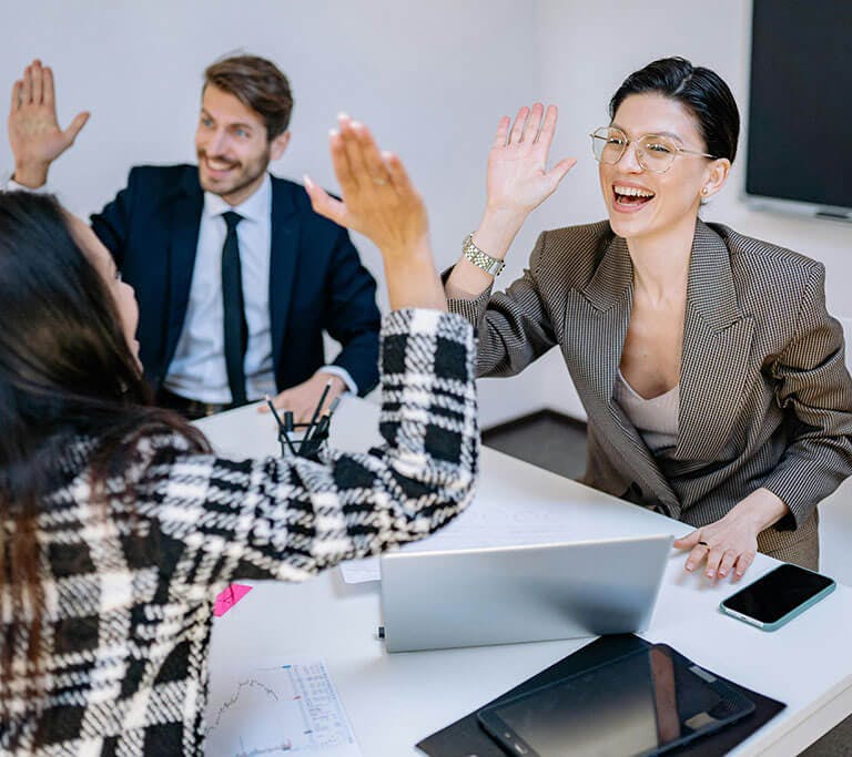 A group of colleagues celebrating success in a meeting by giving high fives around a table with laptops, charts, and office supplies. The atmosphere appears positive and collaborative in a modern office setting.