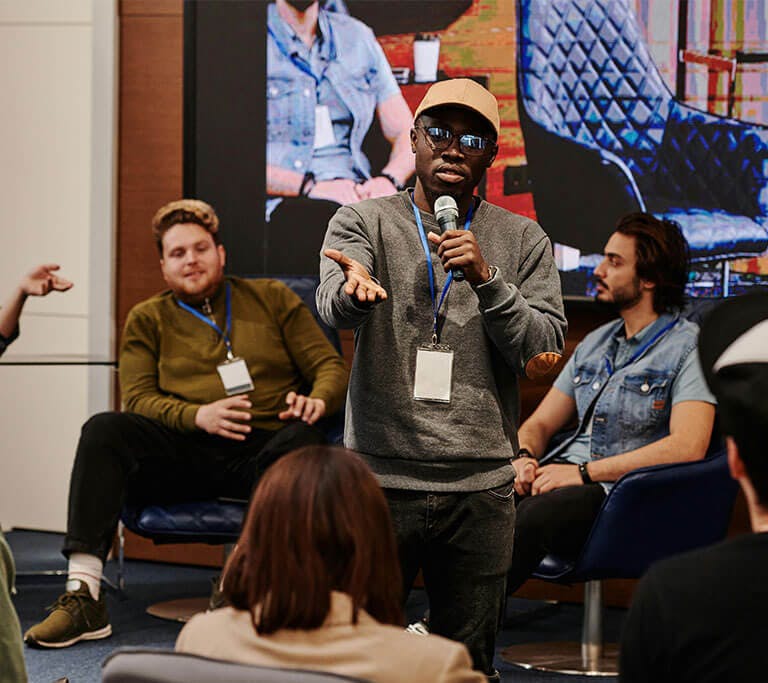 A speaker with a microphone addresses an audience during a panel discussion, with three seated panelists and a large screen in the background.