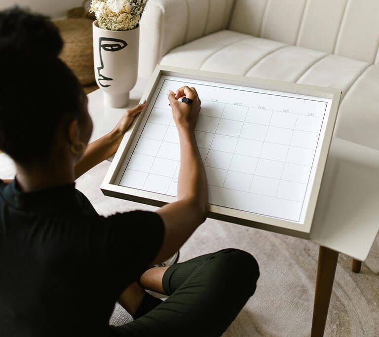 A person sitting on the floor and writing on a blank calendar board placed on a small table beside a white couch in a cozy office.