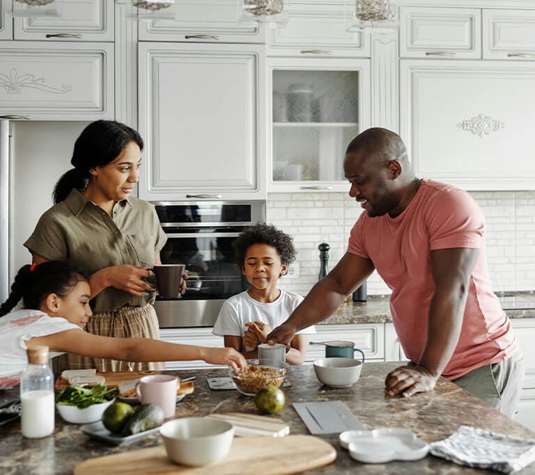 Family stands around a kitchen island preparing and sharing food together, with bowls, fruit and kitchen items spread across the counter.