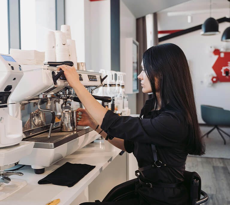 A barista operating a professional espresso machine in a modern café, preparing coffee while surrounded by equipment, paper cups and syrup bottles.
