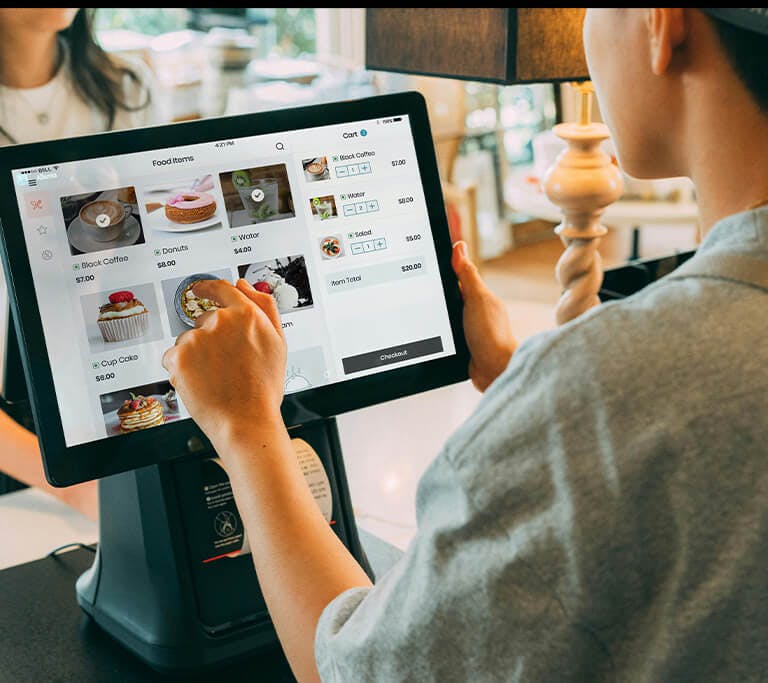 A customer uses a touchscreen point-of-sale system to place an order at a counter, with a worker standing on the opposite side.
