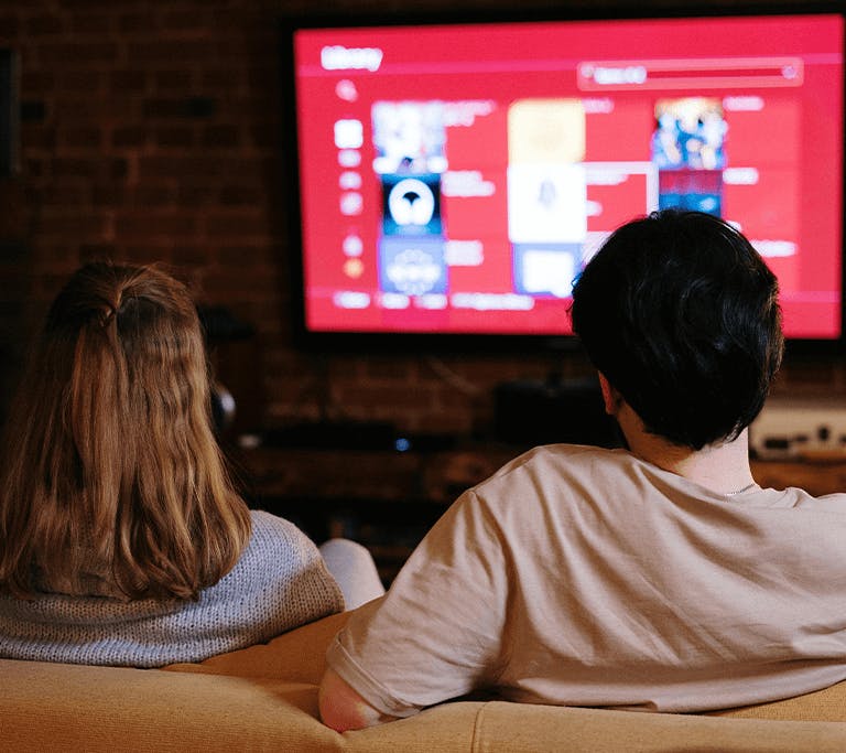 Two people sit side by side on a couch, seen from behind, watching a large television screen with a red interface and various media options displayed in a cozy living room with brick walls.