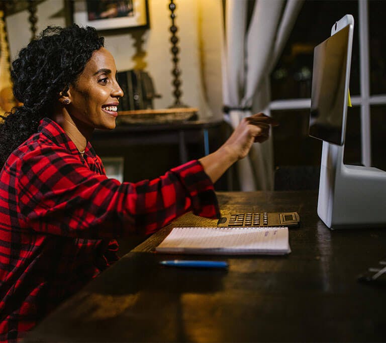 A person with long curly hair wearing a red and black checkered shirt sits at their computer desk while smiling and pointing at a screen.