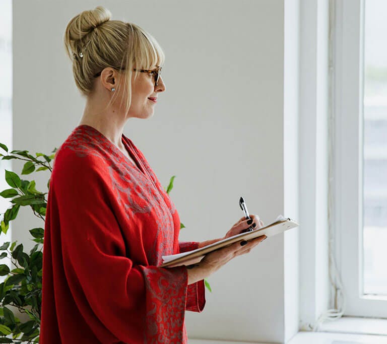 Person in a red patterned robe stands by a bright window, holding a clipboard and pen while looking outside, with green plants in the corner of the room.