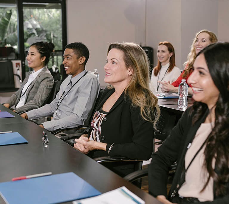 A group of professionals wearing name badges sits in a conference room with folders, pens and water bottles on their desks.