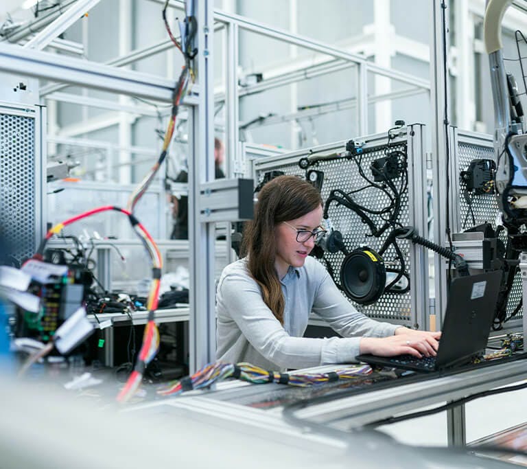A person works on a laptop inside an industrial workspace surrounded by machinery, wiring, and testing equipment.