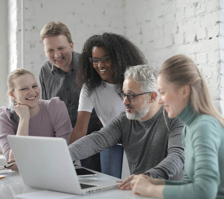 A group of five people gathered around a table, smiling and looking at a laptop screen. One person is pointing at the screen while the others watch and engage in discussion in a bright, modern office with large windows.