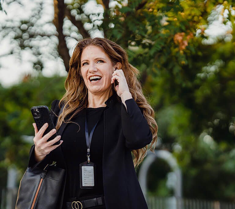A person standing outdoors smiling while holding a smartphone and adjusting an earbud. They are wearing a lanyard and carrying a bag, surrounded by trees and natural light.