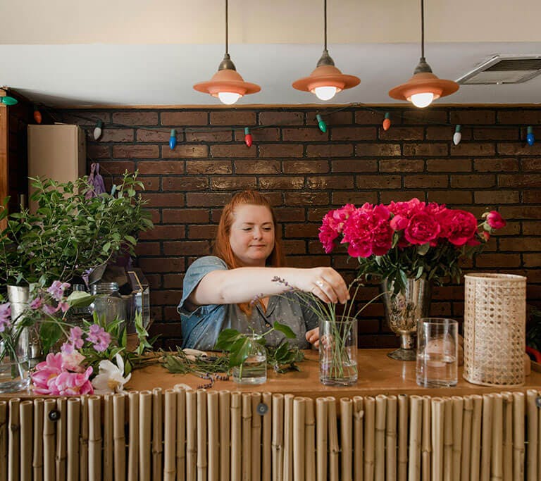 A person arranging flowers at a counter decorated with bamboo, surrounded by colorful blooms and greenery in a cozy space with a brick wall and hanging lights.