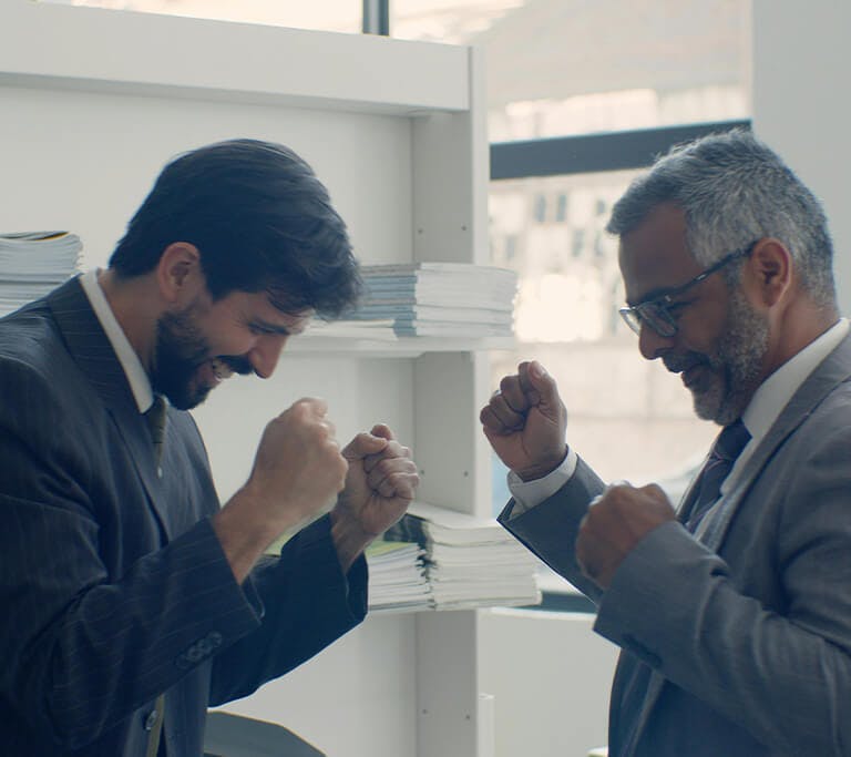 Two businessmen in suits smiling and celebrating with raised fists in an office setting, standing near shelves filled with stacks of documents.