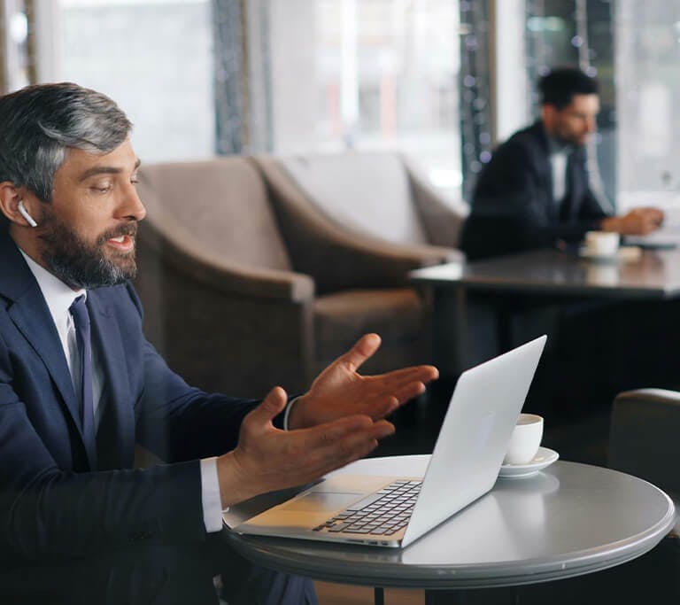A person in a business suit sits at a small table in a café, speaking during a video call on a laptop while using wireless earbuds. In the background, others work and talk at nearby tables.