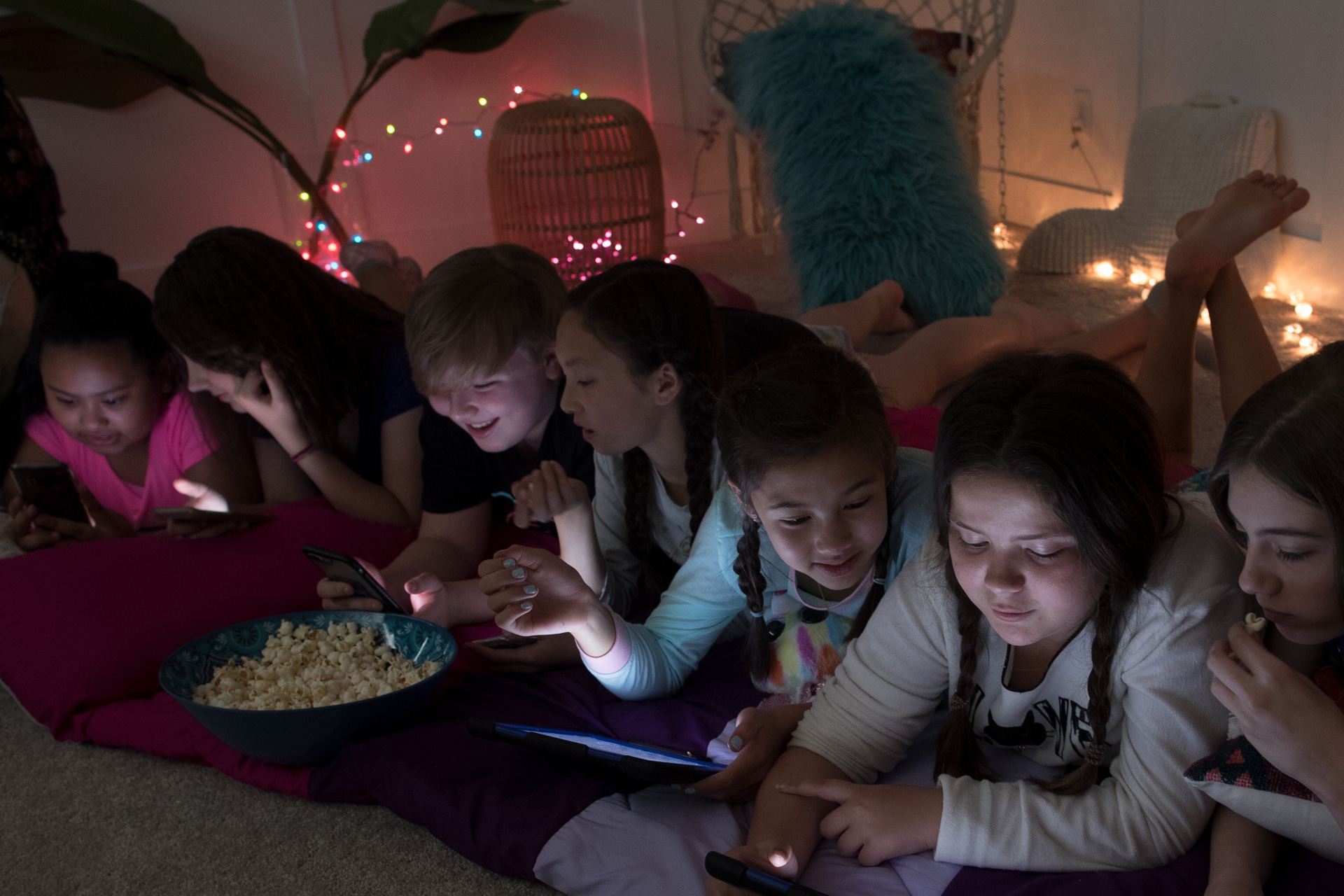 A group of pre-teens girls areat a slumber party. They are all on their devices, laughing and sharing images.